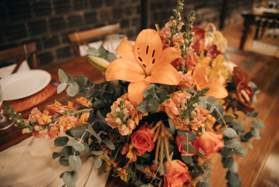 A floral arrangement featuring a vibrant mix of orange lilies with large, open petals and dark stamens, complemented by peach and coral roses with tightly packed petals, mixed with smaller orange and 