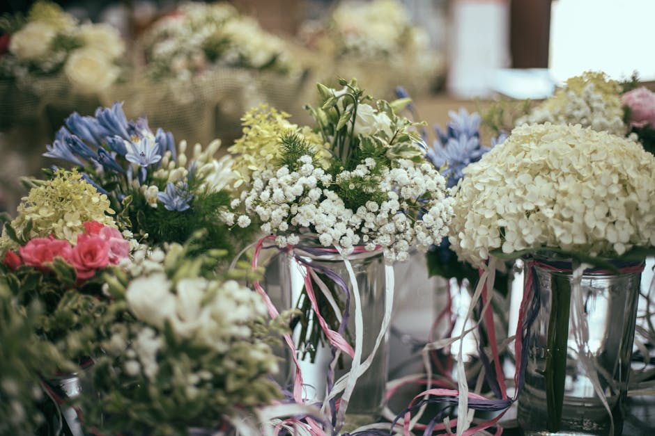 A collection of floral arrangements featuring white hydrangeas, pink roses, blue irises, and white baby's breath, all displayed in clear glass vases decorated with pink and white ribbons. The flowers 