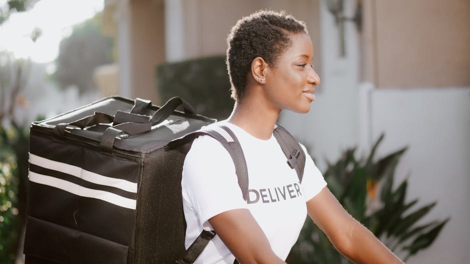 A young woman with short, natural curly hair and a confident expression, dressed in a white T-shirt with the word 'DELIVER' printed on it, is holding a large black insulated delivery bag with white st