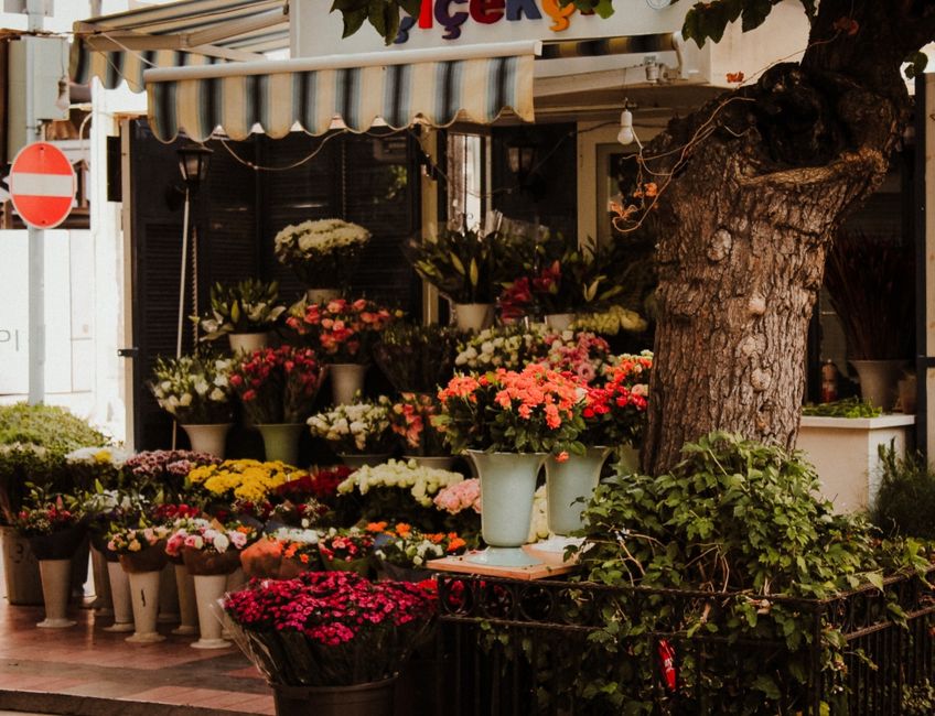 An outdoor flower shop displaying a vibrant variety of fresh flowers arranged in white and black pots, including pink, red, yellow, and white blooms such as chrysanthemums and roses. The arrangement i
