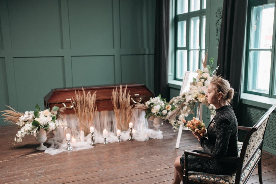 A woman seated indoors on an ornate wooden chair, holding a bouquet of peach and cream-colored flowers, with soft natural light streaming through large green-framed windows behind her. In front of her