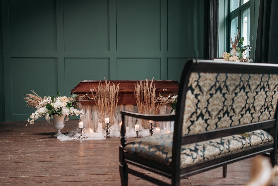 A funeral setting with an arrangement of flowers placed on a wooden table in front of a closed wooden casket. The floral display includes white roses, lilies, and greenery in a white ceramic vase, com